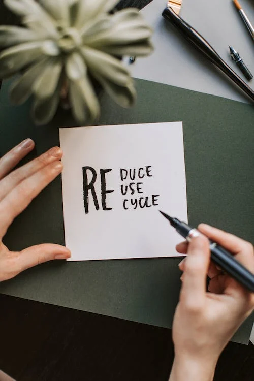 Person writing on a paper with the text "Reduce Reuse Recycle" using a black marker, next to a green plant and office supplies on a green desk.