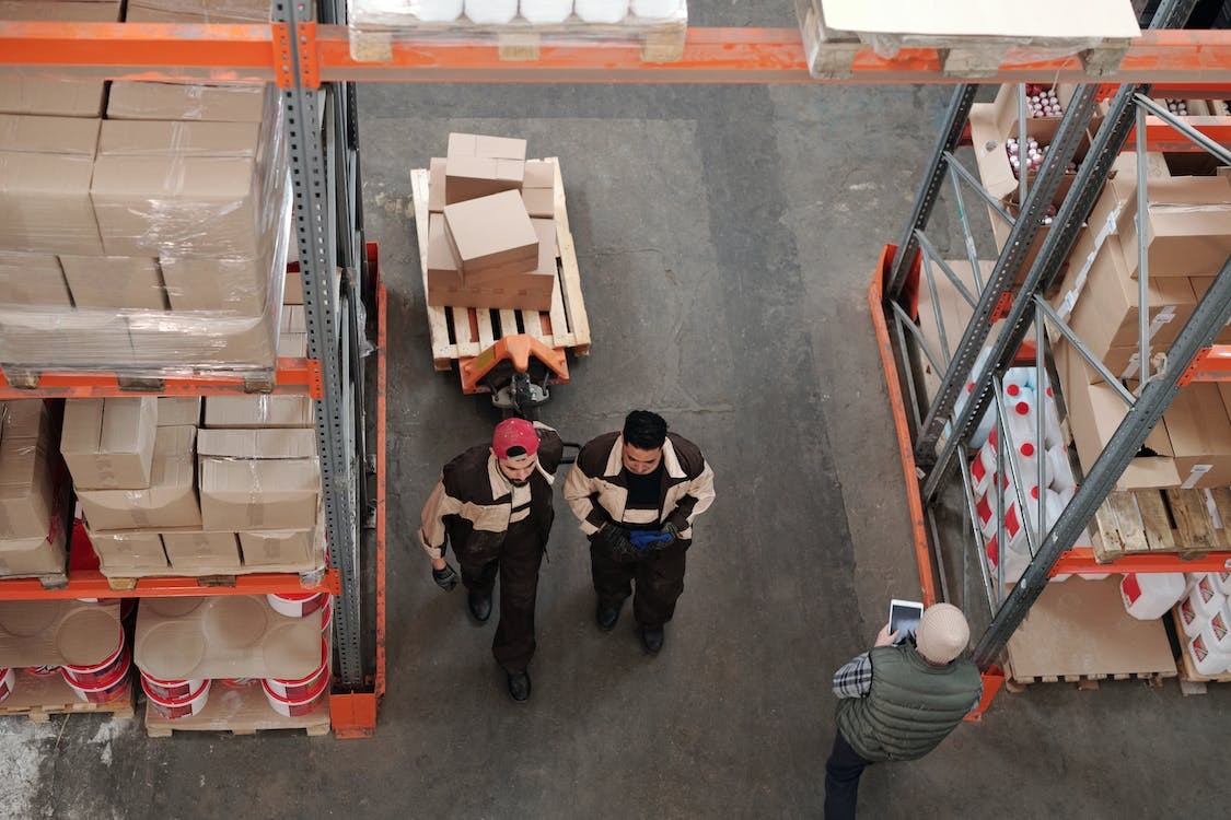 Warehouse workers in uniform walking through a storage area, transporting cardboard boxes on a pallet jack, with shelves of organized inventory in the background.