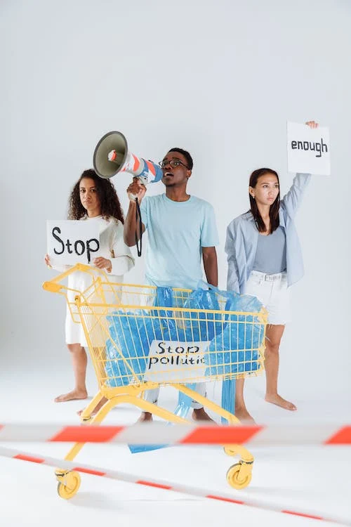 Three people protesting with signs that say "Stop" and "Enough," one holding a megaphone, standing behind a shopping cart filled with plastic bags and a sign that says "Stop Pollution."