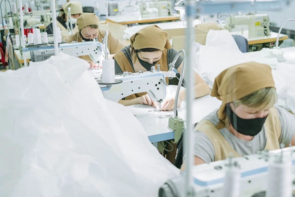 Factory workers wearing masks and headscarves operating sewing machines in a textile manufacturing facility.