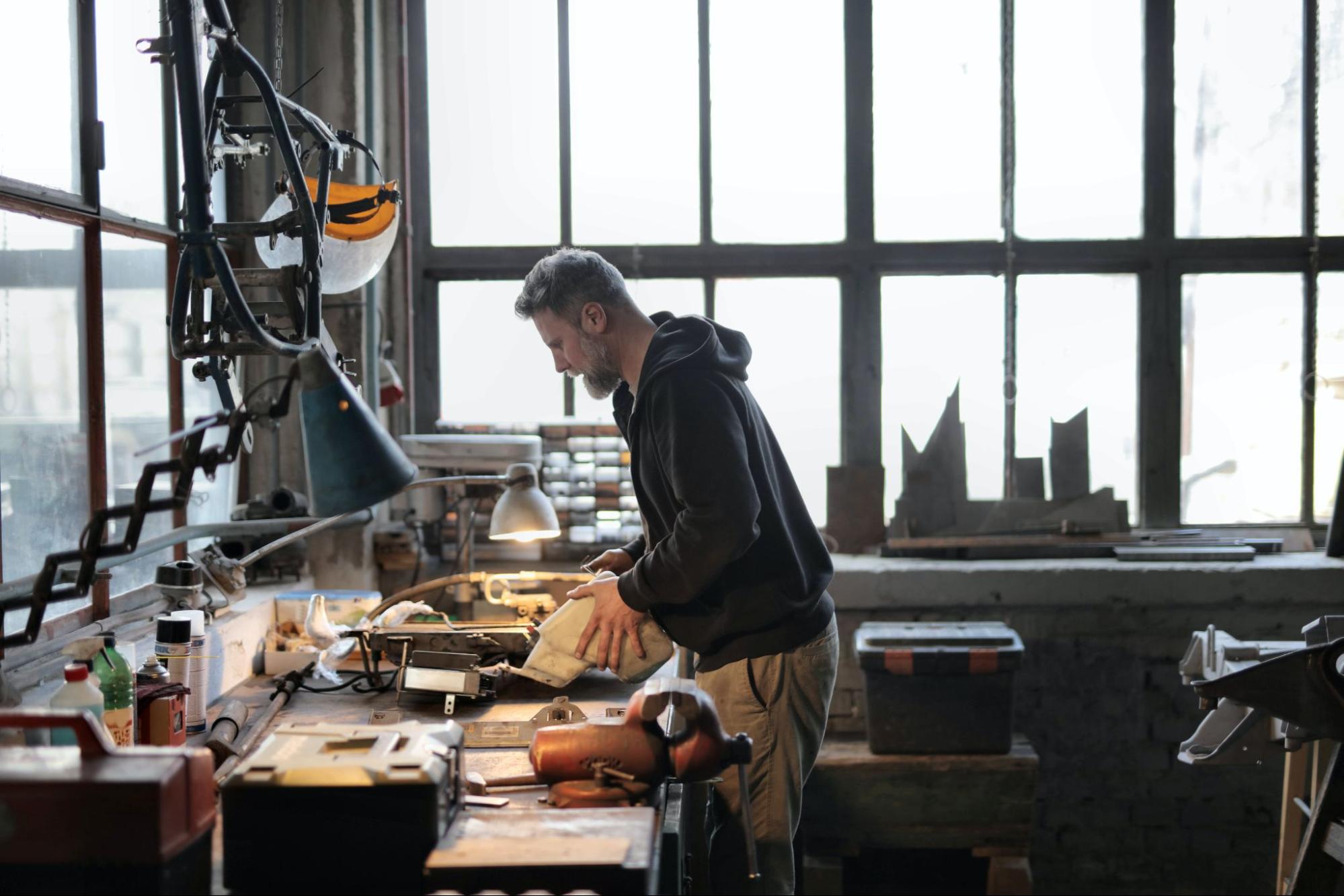 Man in black hoodie holding object in workshop with assorted tools on table