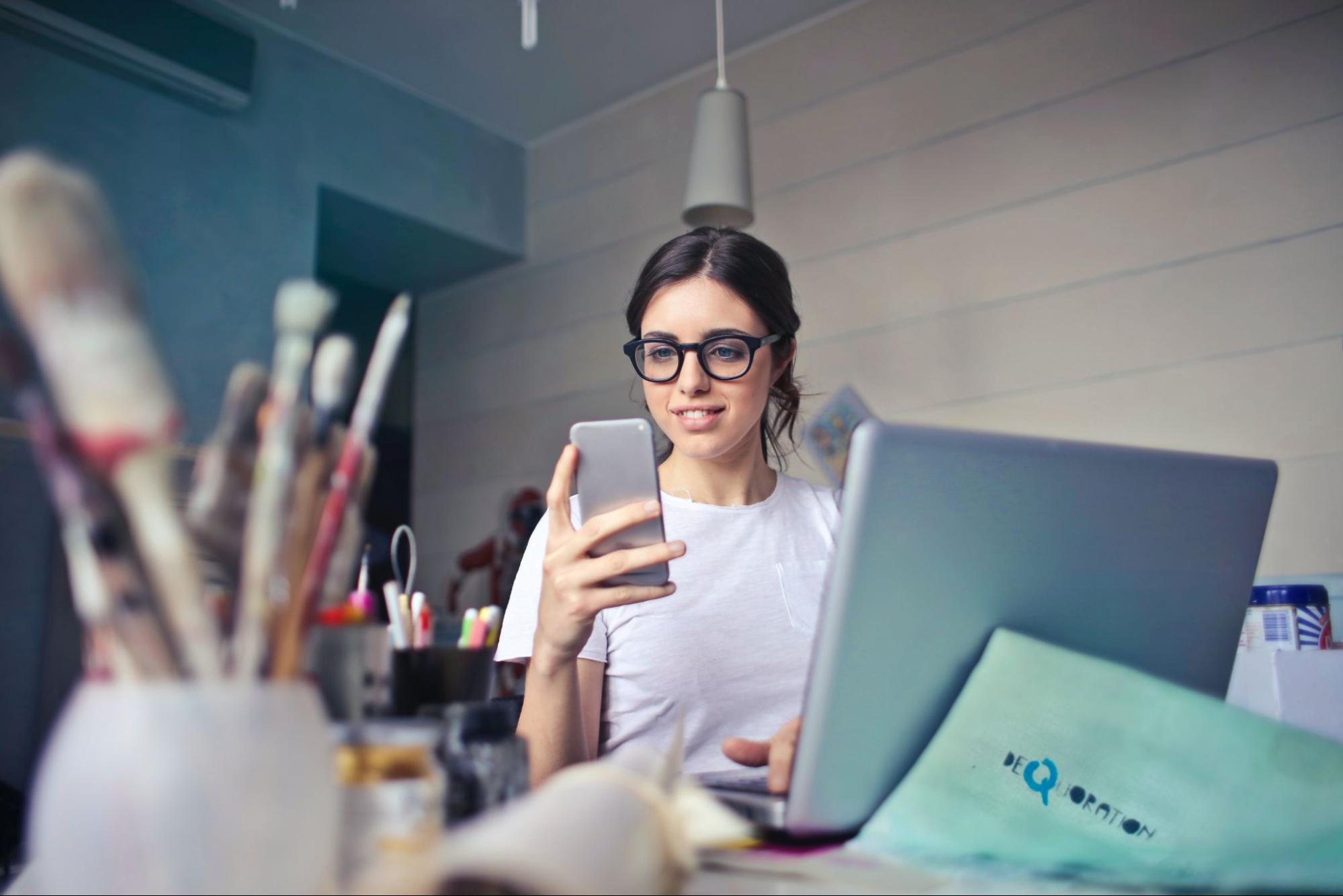 Woman in white shirt and eyeglasses using phone at desk with laptop and art materials